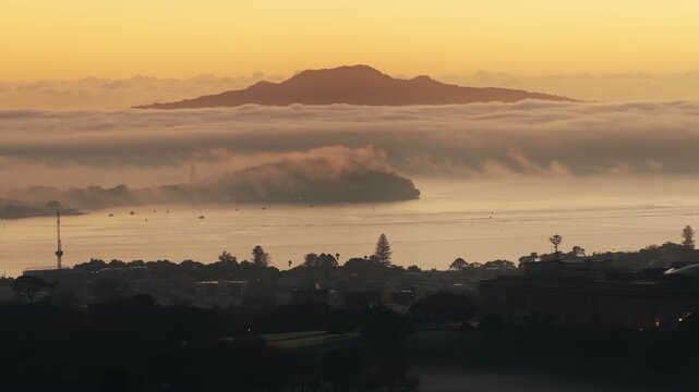 Auckland, New Zealand. Sunrise over the Waitemata Harbour with Rangitoto Island shrouded in mist. Boats are anchored in the calm waters below the city skyline.