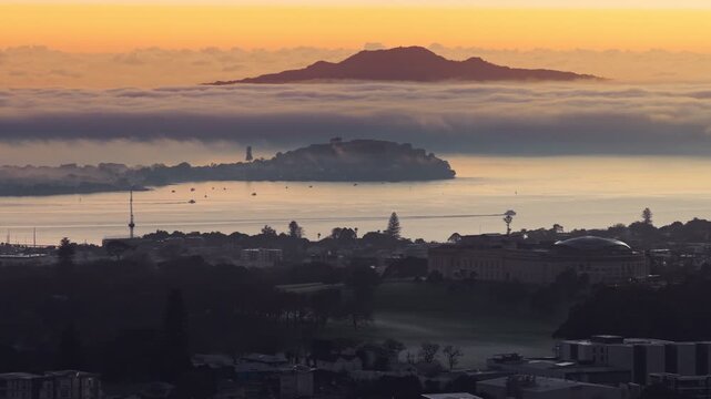 Auckland, New Zealand. Sunrise over the Hauraki Gulf, with Rangitoto Island emerging from the clouds. Boats dot the calm water below the misty city skyline.