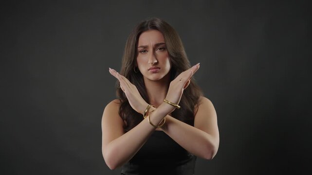 Rejection and disapproval concept with a young woman making an X shape with her arms to signal no or stop. This studio shot captures her serious expression and nonverbal communication. The gesture ref