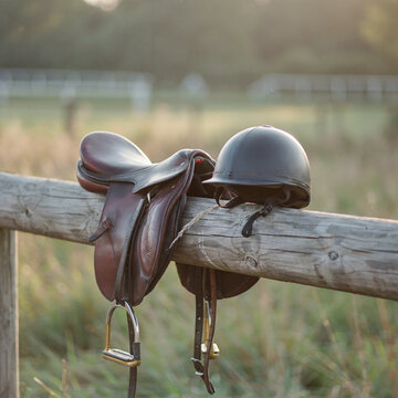 Horse saddle and a protective riding helmet rest balanced together upon a wooden fence rail at sunset outdoors.