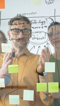 Business people colleagues brainstorming, writing strategy notes on glass board, collaborating with colorful sticky notes in creative workplace meeting in bright office