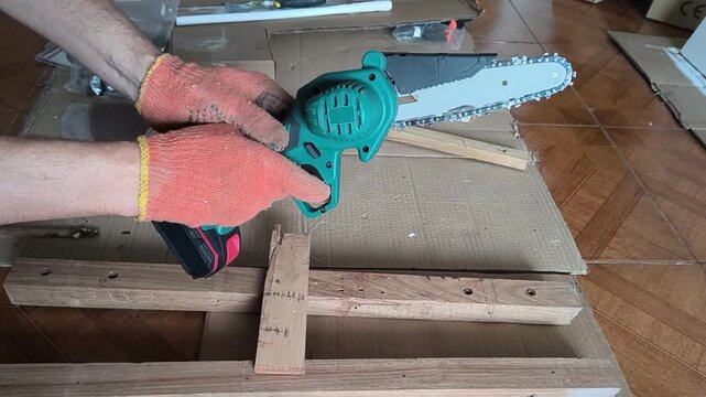 Skilled worker wearing protective orange gloves holds a small cordless electric chainsaw over wood planks in a workshop, ready for a precise DIY carpentry or home repair project.