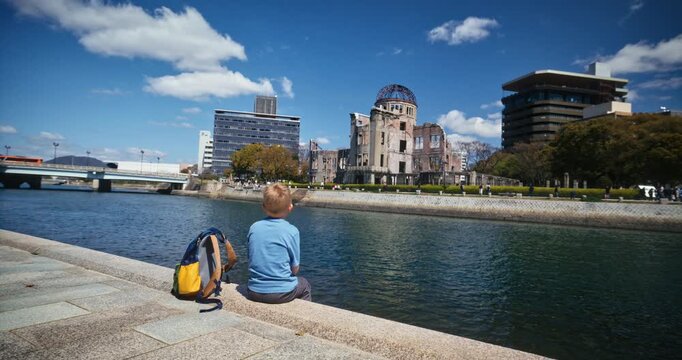 Child With Backpack Sitting at Canal Edge Looking at Hiroshima Peace Memorial Atomic Bomb Dome - Wide Shot