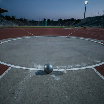 Shot put ball sits alone in the center of the concrete throwing circle on a large stadium field at dusk time.