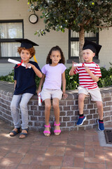 Three children sitting on low brick planter in courtyard wearing caps and holding red diplomas © wavebreak3