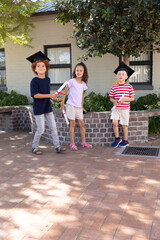 Diverse children wearing mortarboards holding red-ribboned diplomas on brick courtyard celebrating © wavebreak3