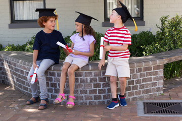Three children sitting on curved planter at school wearing black caps, holding red-ribbon diplomas © wavebreak3