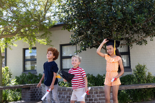 Diverse children holding black caps with yellow tassels and rolled certs, red ribbons in front yard