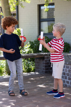 Diverse boys exchanging rolled white scrolls tied with red ribbons on paved brick patio near window