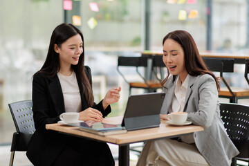 Obraz premium Two smiling asian businesswomen engaging in a lively professional discussion, exchanging ideas and collaborating on projects while sitting at a table with coffee cups and a tablet