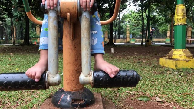 Barefoot child in blue pajamas playing on outdoor gym equipment at a public park surrounded by green trees