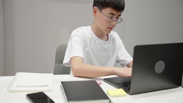 Focused teenage student in glasses using laptop at home desk. Modern education, remote learning, homework, digital study and online school technology concept.