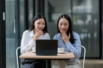Obraz premium Two smiling asian businesswomen sharing a tablet, covering their mouths in surprise and laughter during a casual meeting or coffee break, highlighting collaboration and digital connection
