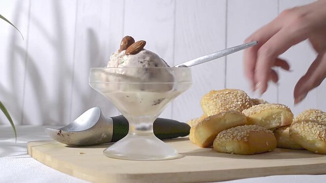 Vanilla ice cream with chocolate in a glass cup, with almonds on top, illuminated by window light on a summer afternoon and a rustic white wooden wall in the background.  putting spoon