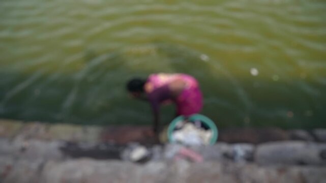 Blurry, high-angle view of a poor indian woman in a traditional sari washing clothes by hand on the stone steps of a polluted river, representing daily life and poverty in developing countries