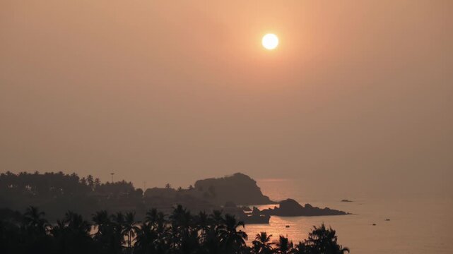 Tranquil sunset casting a warm, golden glow over the hazy sky, with the sun reflecting on the calm ocean water next to a rocky coast silhouetted with lush palm trees