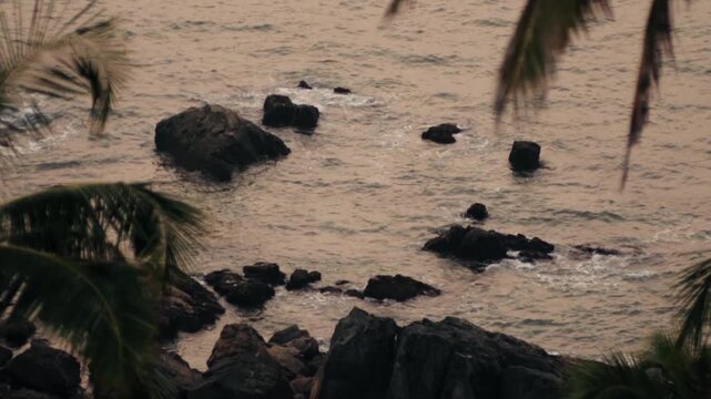 Scenic view of a rocky tropical seashore with gentle ocean waves crashing against the cliffs and palm tree fronds swaying in the foreground, creating a tranquil and beautiful natural landscape