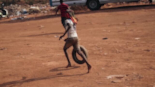 Barefoot african child with dreadlocks playing and having fun, running on a dusty road while pushing an old tire with a stick in a poor village, showing the simple joys of childhood