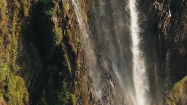 White water rushing down a steep, rocky cliff face covered with green moss and vegetation in a sunlit natural environment, showcasing the immense power and beauty of a wild cascade