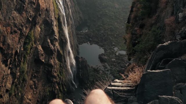 Point of view shot of a brave adventurer sitting on the edge of a high cliff, dangling his feet over the abyss while enjoying the stunning view of a waterfall plunging into a lake below