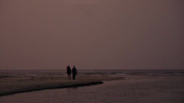 Silhouetted couple walking together along a deserted and moody shoreline during a tranquil and melancholic sunset, enjoying a peaceful and romantic moment in a vast, empty natural landscape