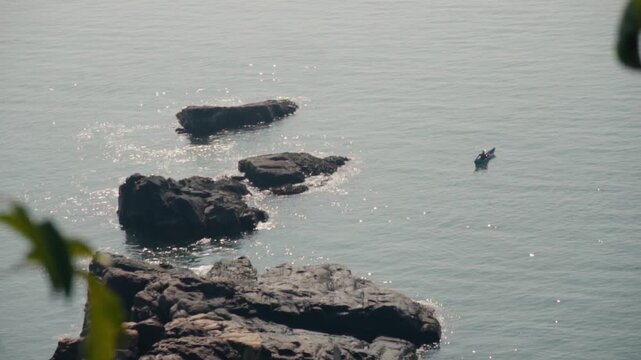 Aerial view of a solitary fisherman navigating a traditional wooden boat across the tranquil, sun-dappled sea, passing by a series of rugged rock formations on a peaceful morning