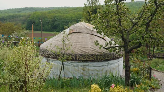 Authentic nomadic yurt standing in a verdant garden with blooming flowers and trees, set against a backdrop of distant green hills, representing a traditional and eco-friendly lifestyle