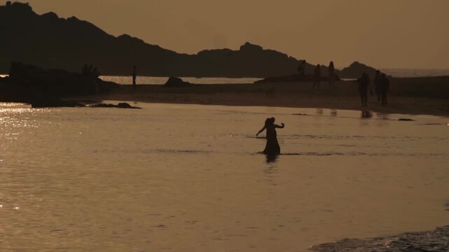 Silhouettes of two people playing and splashing in the calm sea during a beautiful golden sunset, with other tourists walking on the sandy beach and rocky mountains in the background