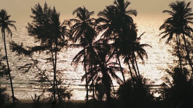 Dramatic silhouette of palm trees swaying in the wind against a sparkling sea during a golden sunset, with unrecognizable people strolling along the shoreline in the background