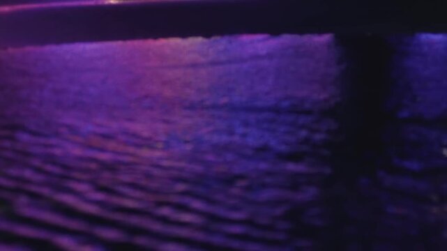 Mesmerizing dark water surface reflecting vibrant purple, blue, and pink city lights at night, creating a beautiful and tranquil shimmering ripple effect with a cinematic mood