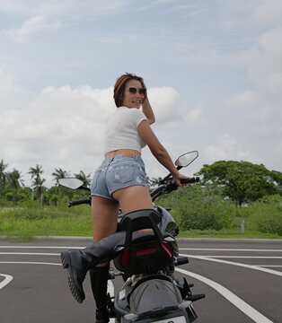 young Asian woman in denim shorts and white crop top climbs onto a motorcycle on an asphalt road with tropical trees in the background