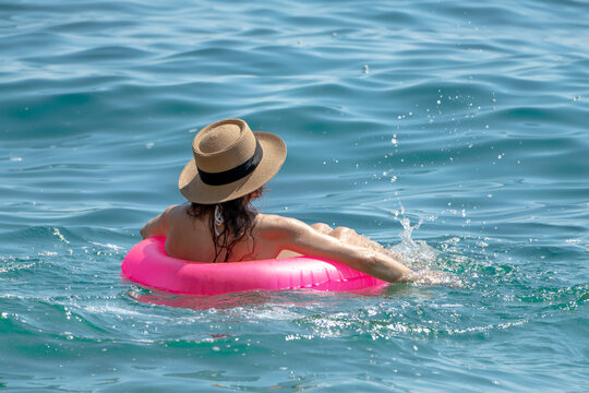 Woman Swimming Sea: Woman in hat relaxing, floating pink tube, splashing warm ocean water on sunny day.