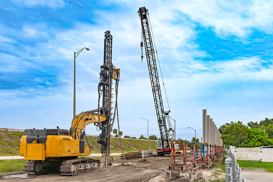 Heavy drilling rig and lattice boom crawler crane installing concrete sound barrier wall panels on highway construction site