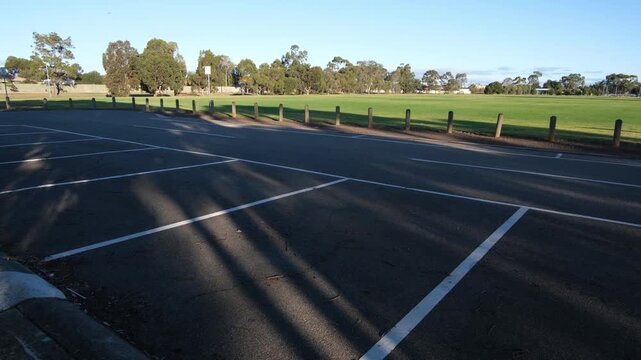 An empty asphalt car park with white painted bay markings sits adjacent to a large green oval and open parkland in an Australian suburban.&nbsp;Timber bollards line the boundary of the sports ground