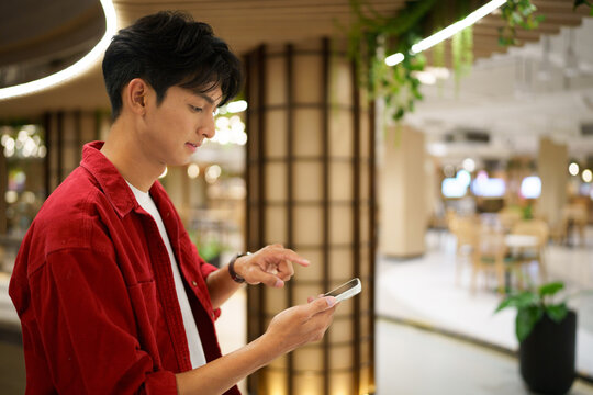 Young Asian Man Using Smartphone in Modern Shopping Mall with Bokeh Background