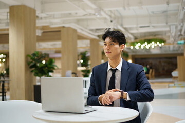 Fototapeta premium Young Asian Businessman Sitting at Table with Laptop in Modern Co working Space Looking Thoughtful