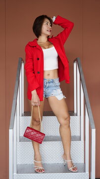 Smiling woman wearing a red coat over a white crop top and denim shorts stands on metal stairs
