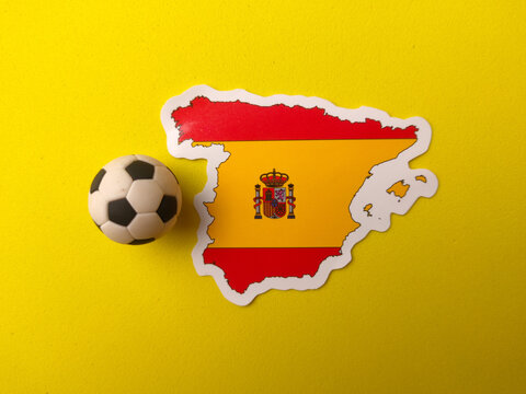 A Spain map sticker printed with its national flag design sits next to a miniature football on a yellow background