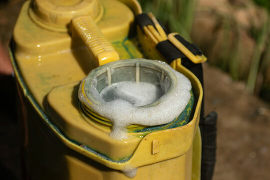 White chemical foam bubbling up around the filter mesh of a yellow agricultural spray tank during mixing process
