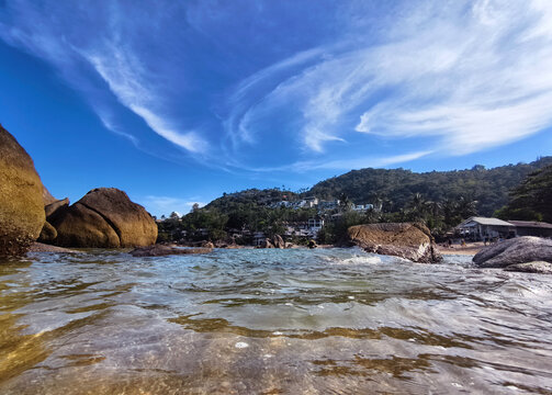 Rocky tropical beach clear turquoise water, sweeping cloud formations, sunlit boulders in shallow.
