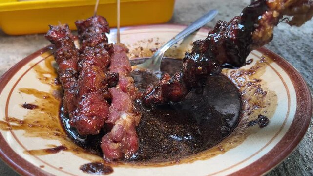 close up view of goat satay being coated with spices on the plate. skewers. eid al adha. idul adha. sate kambing, sapi. indonesian food or culinary. side, front. stainless steel, spoon.