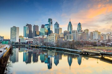 Naklejka premium Philadelphia skyline reflected in the Schuylkill River during sunrise, featuring iconic Center City skyscrapers, calm water, soft pastel clouds, and a tranquil urban waterfront scene.