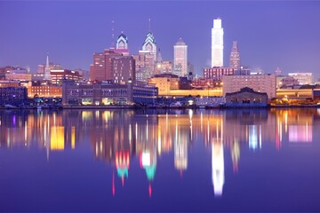 Fototapeta premium Philadelphia skyline illuminated at night across a calm waterfront, featuring glowing skyscrapers, colorful city lights, and vivid reflections creating a dramatic urban panorama.