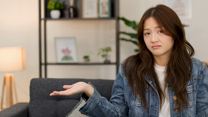 Fototapeta premium Confused Young Asian Woman Shrugging Indoors on a Living Room Sofa