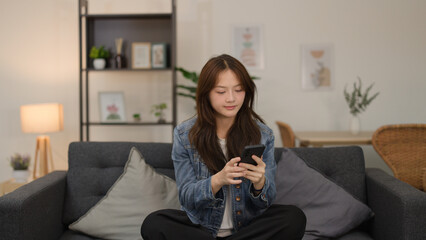 Obraz premium Young Asian Woman in Denim Jacket Using Smartphone on Living Room Sofa