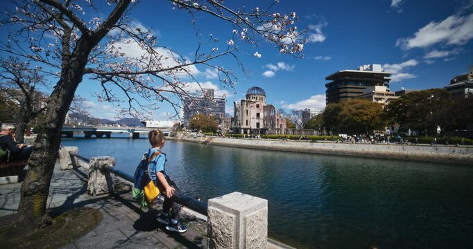 Child Sits On rail of river's edge and looks across at Hiroshima peace memorial dome - wide shot
