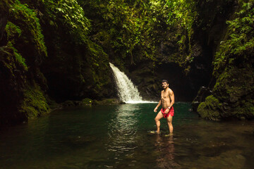 Fototapeta premium Vertical landscape shot with an adult male tourist enjoying himself in front of a waterfall with a natural pool of fresh, pure water in the middle of a tropical forest in Costa Rica