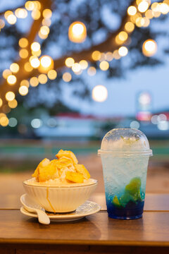 Delicious milk bingsu shaved ice topped with crispy butter toast cubes served with a blue Italian soda cocktail on a wooden table with gorgeous night bokeh lights.