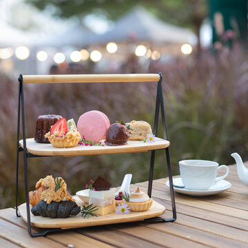 Close-up of an elegant two-tier wooden afternoon tea stand filled with assorted gourmet desserts and pastries on a wooden table at an outdoor cafe.