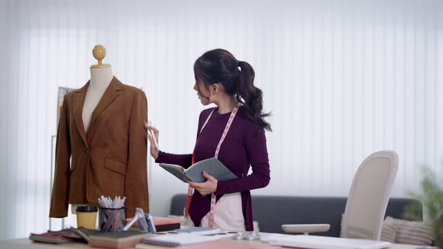 Young Asian fashion designer examining brown blazer on mannequin while holding sketchbook and measuring tape in bright modern studio workspace